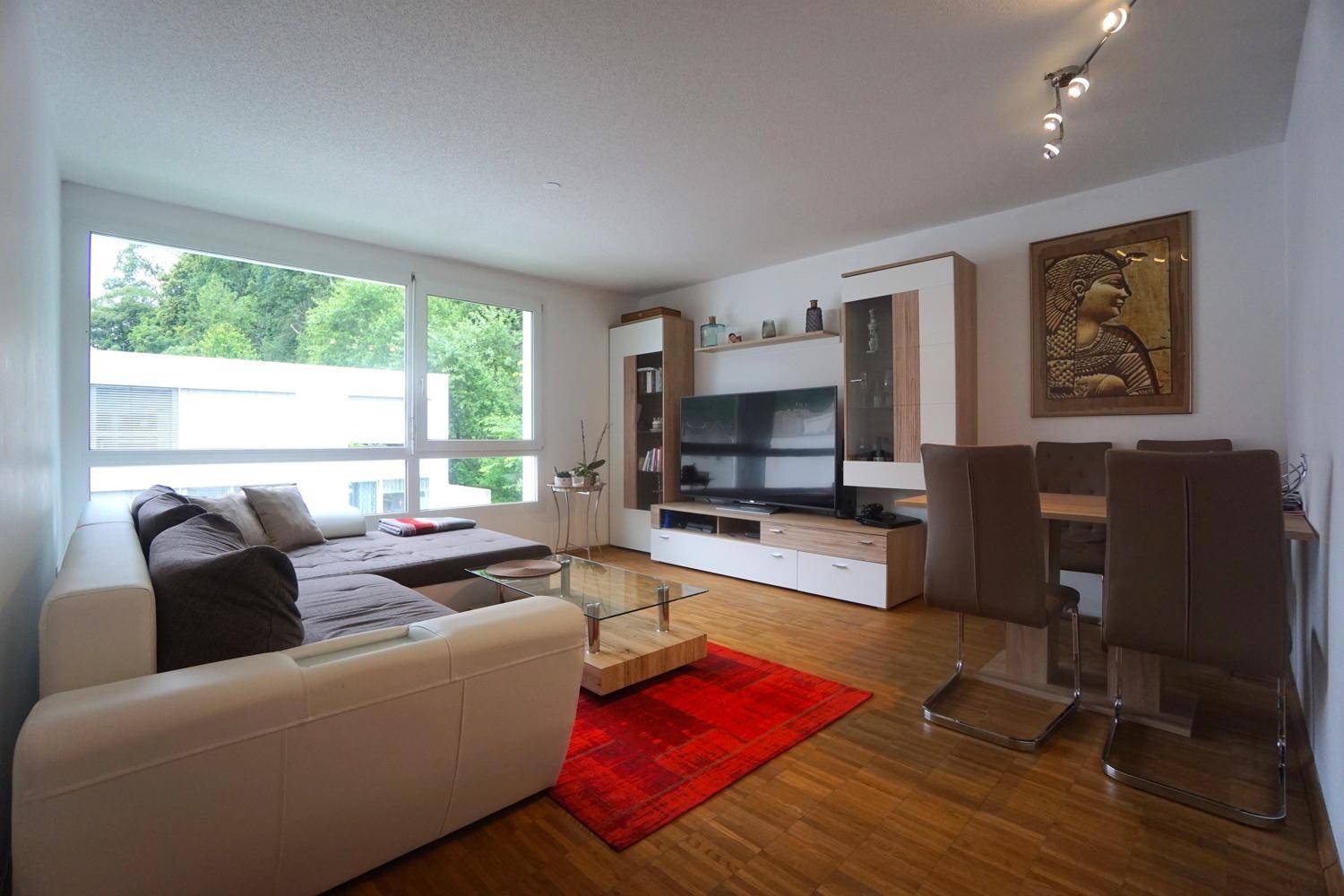 Geräumiger Wohn-Essbereich mit bequemer Couch, Esstisch und viel Tageslicht durch große Fenster.