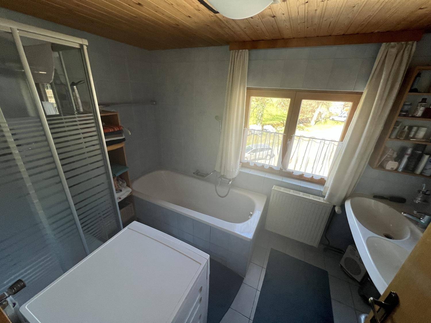 Gefliestes Badezimmer mit Badewanne, Duschkabine, Waschbecken und Fenster mit Blick ins Grüne.