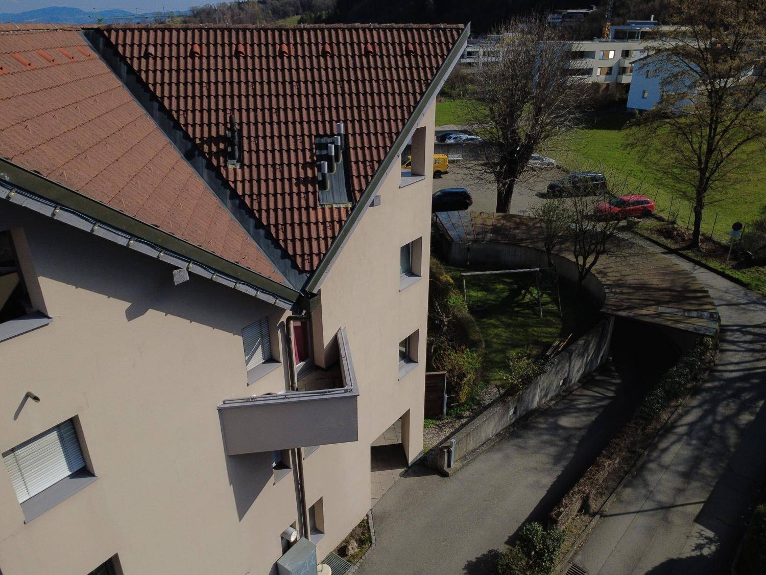 Blick auf die Seitenfassade eines Wohnhauses mit Ziegeldach und Balkon, eingebettet in eine grüne Umgebung.