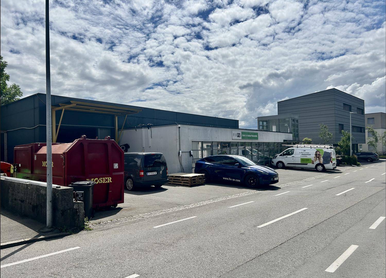 Gewerbegebäude mit Parkplätzen vor der Fassade und einem roten Container auf der linken Seite.
