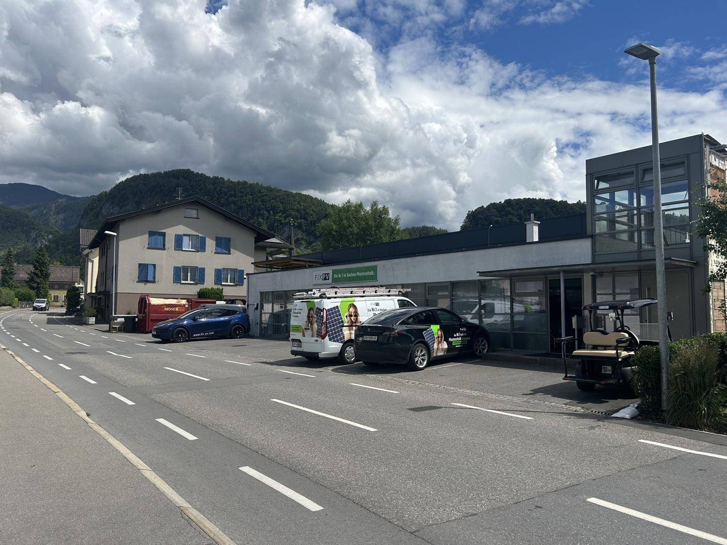 Gewerbegebäude an einer Straße mit geparkten Autos und einem traditionellen Haus im Hintergrund vor Bergkulisse.