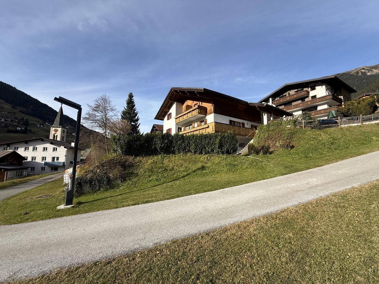 Traditionelles Haus am Hang mit Holzbalkonen und einem Kirchturm im Hintergrund unter blauem Himmel.