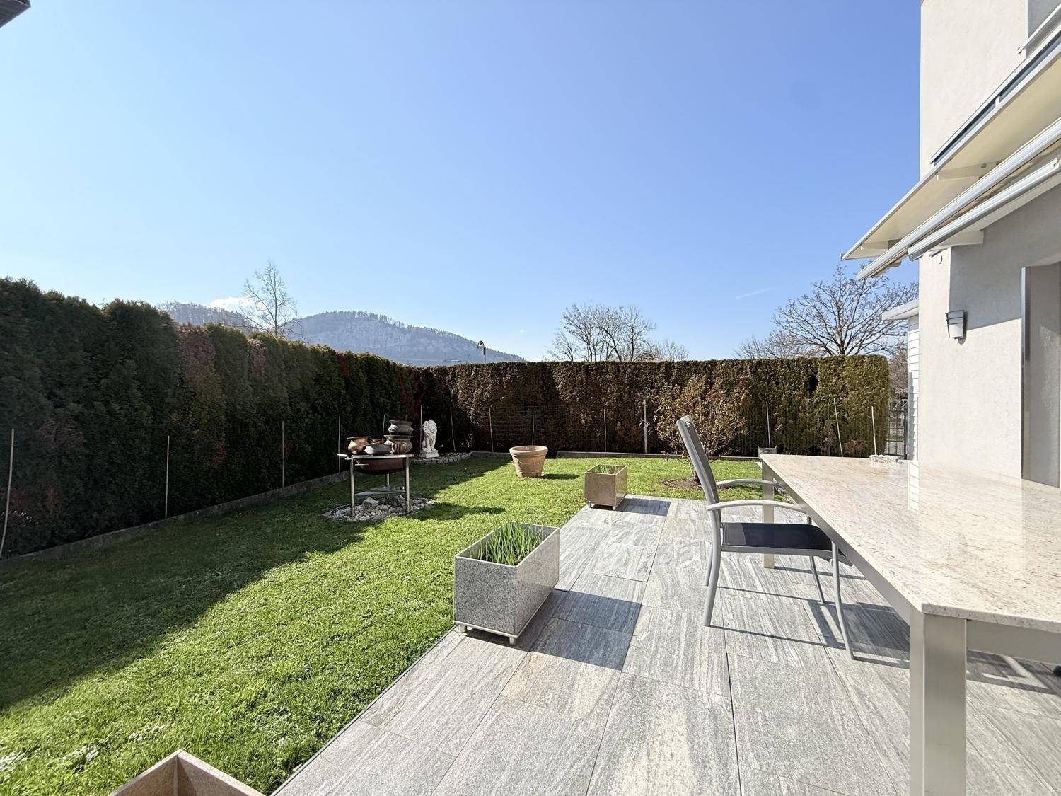 Sonnige Terrasse mit Gartenmöbeln, Grillplatz und hohem Heckenbewuchs für Privatsphäre.