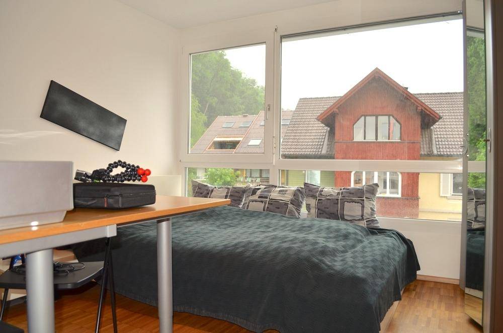 Schlafzimmer mit Holzboden, großem Fenster und Blick auf die umliegenden Gebäude, ausgestattet mit Bett und Schreibtisch.