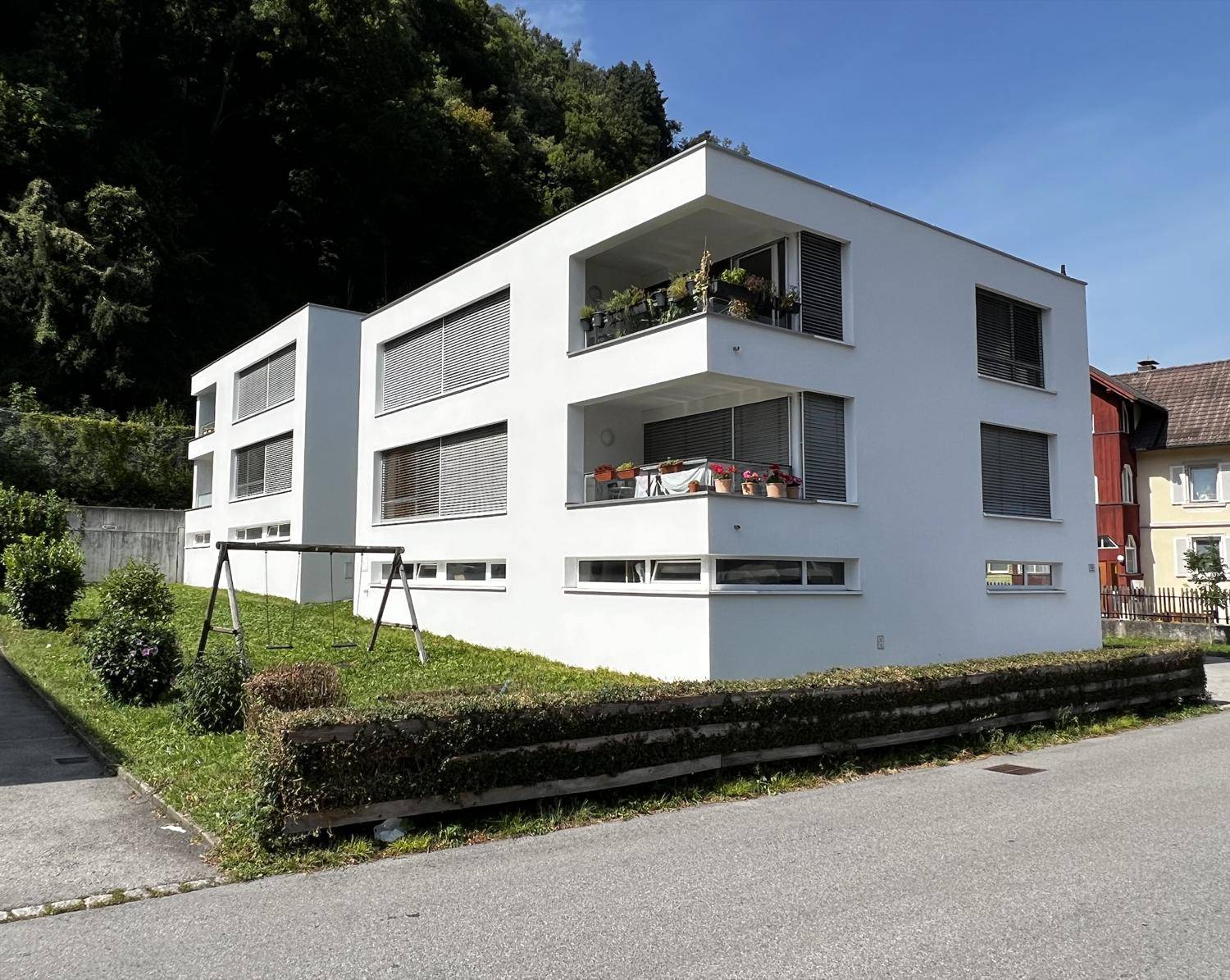 Ansicht der modernen weißen Wohngebäude mit Balkonen, großen Fenstern und Jalousien in einer grünen Umgebung.
