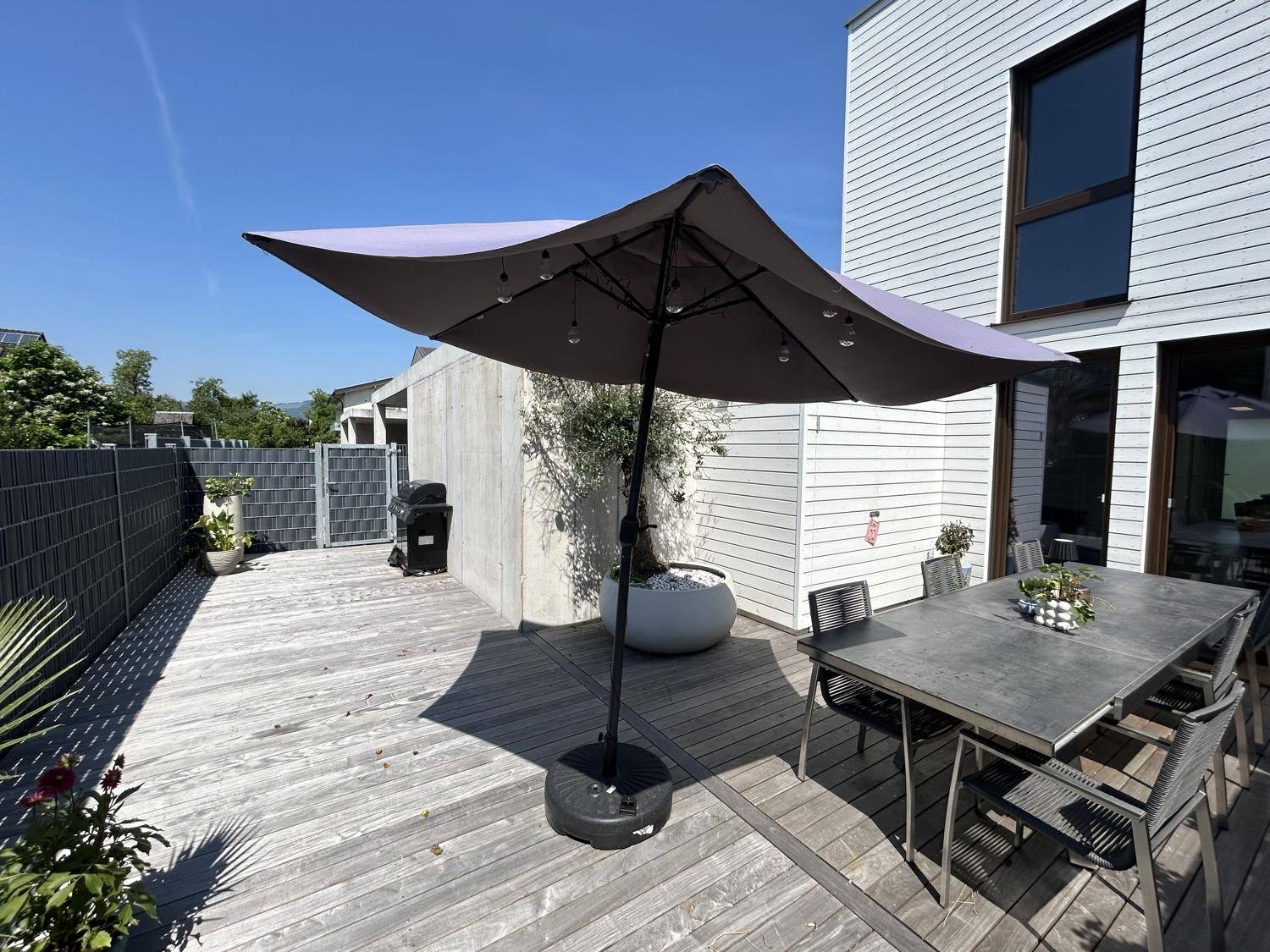 Geräumige Holzterrasse mit Esstisch, Sonnenschirm und Blick ins Grüne, ideal für Mahlzeiten im Freien.