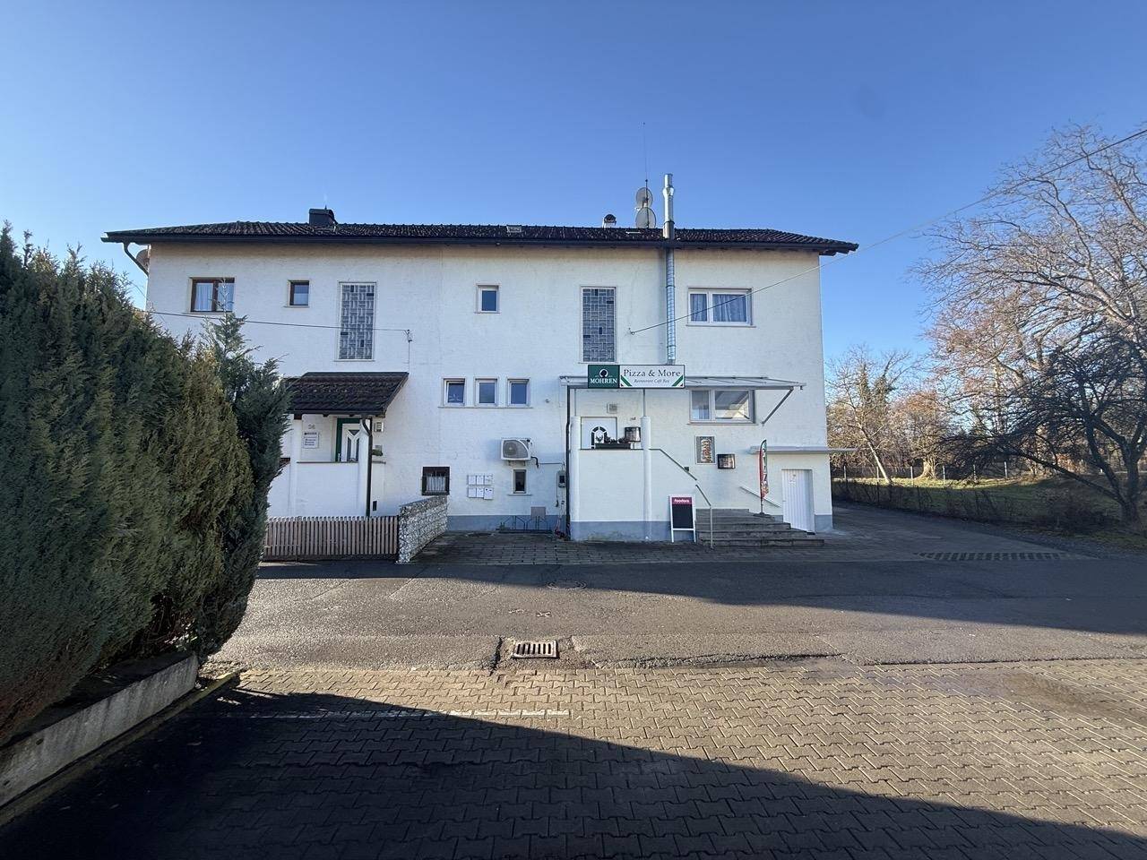 Außenansicht eines weißen Gebäudes mit gepflastertem Vorplatz unter blauem Himmel.