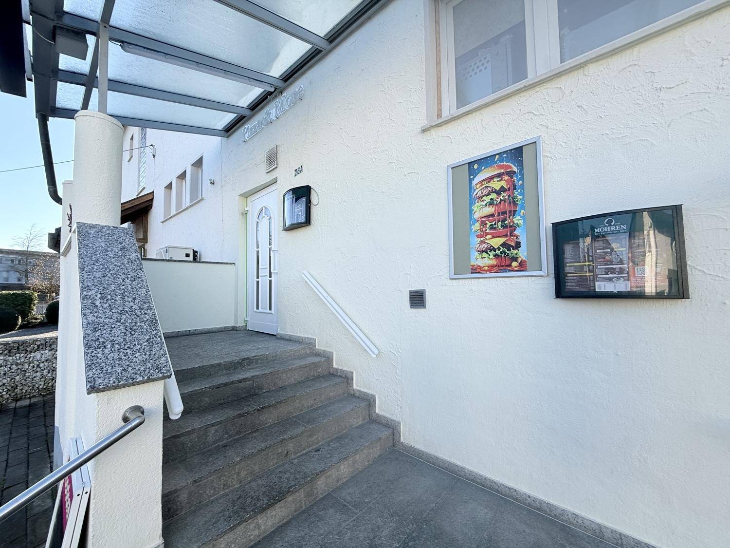 Eingangsbereich des Gebäudes mit Treppe, Überdachung und Werbeplakaten.