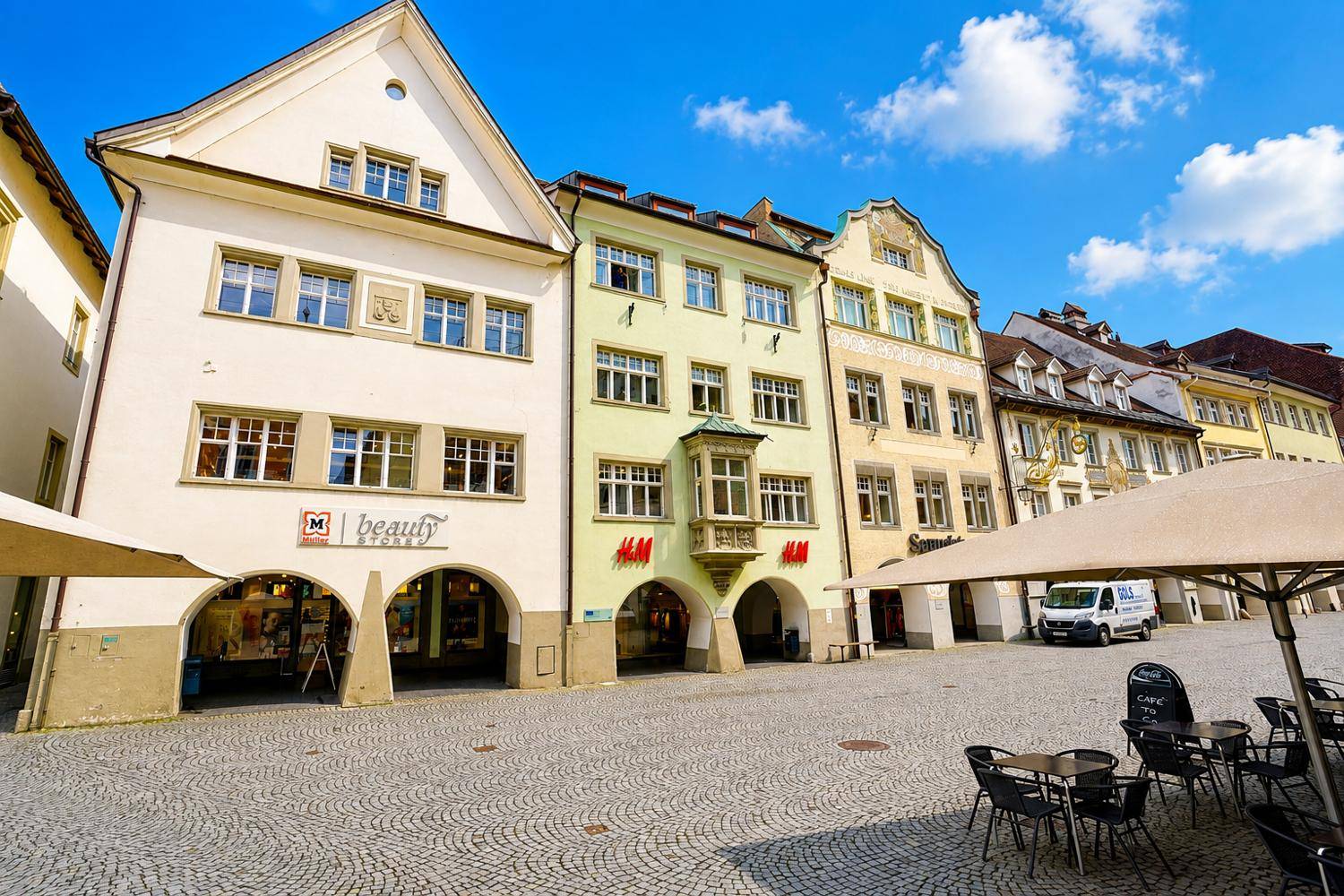 Historische Gebäude an einem belebten Stadtplatz mit Außengastronomie und blauer Himmel.