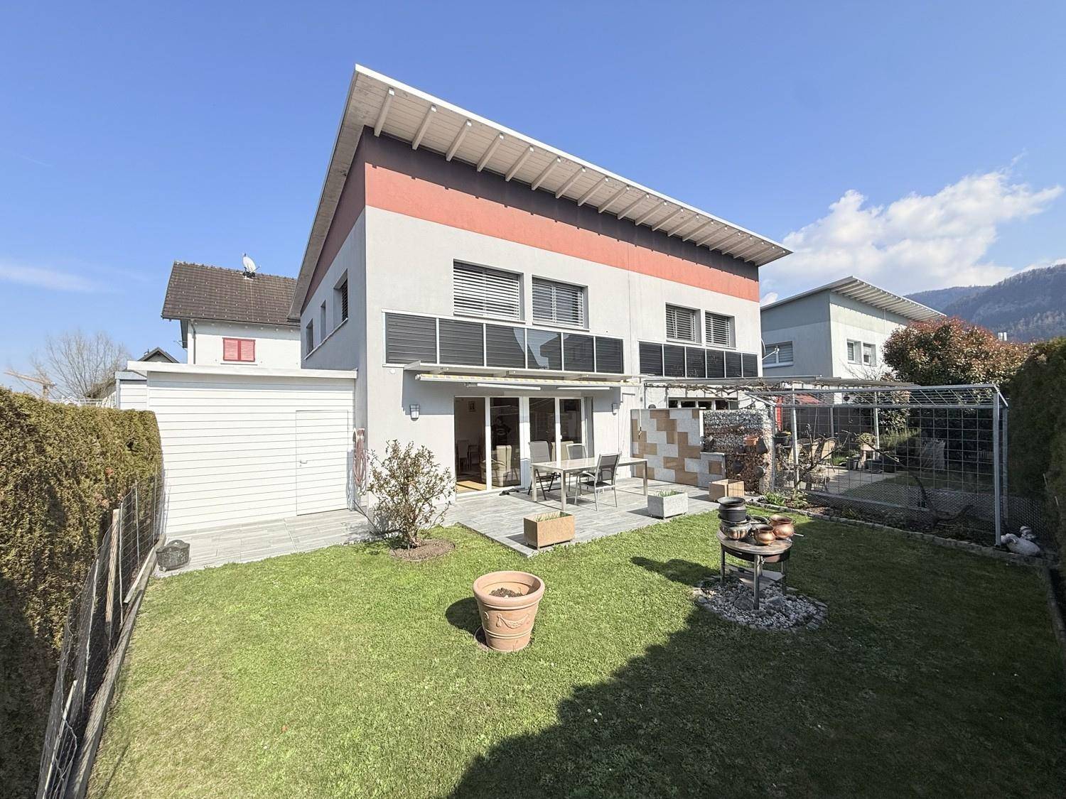Rückansicht des Hauses mit sonnigem Garten, gepflasterter Terrasse und Bergblick.