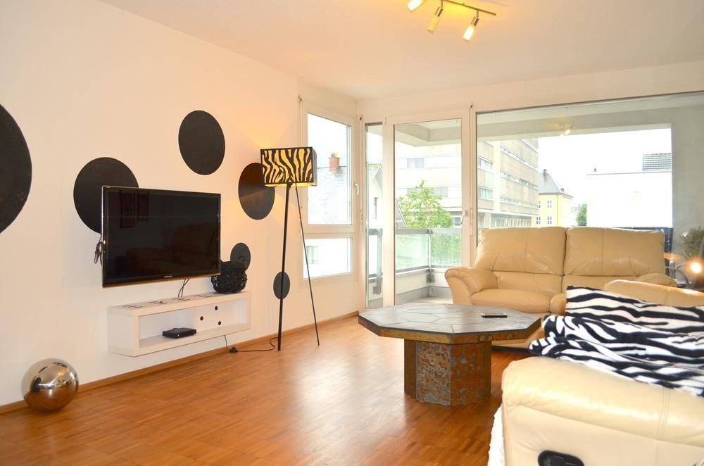 Helles Wohnzimmer mit beigem Ledersofa, Holzboden und Zugang zu einem Balkon, dekoriert mit schwarzen Punkten.