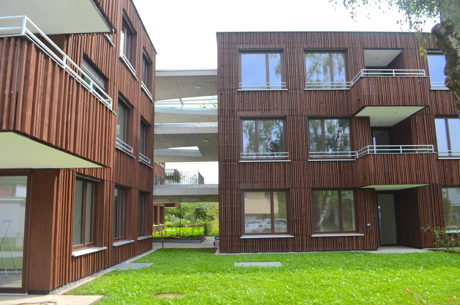 Ansicht des modernen Gebäudekomplexes mit Holzfassaden, verbundenen Flügeln und gepflegter Rasenfläche.
