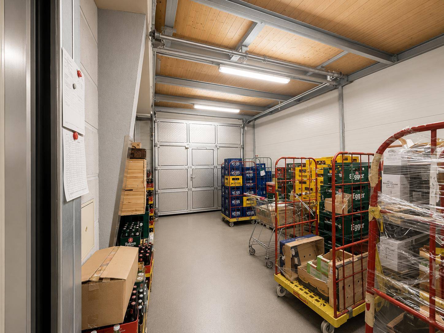 Lagerbereich mit einem großen Rolltor, gestapelten Kisten und Rollcontainern unter einer Holzdecke.
