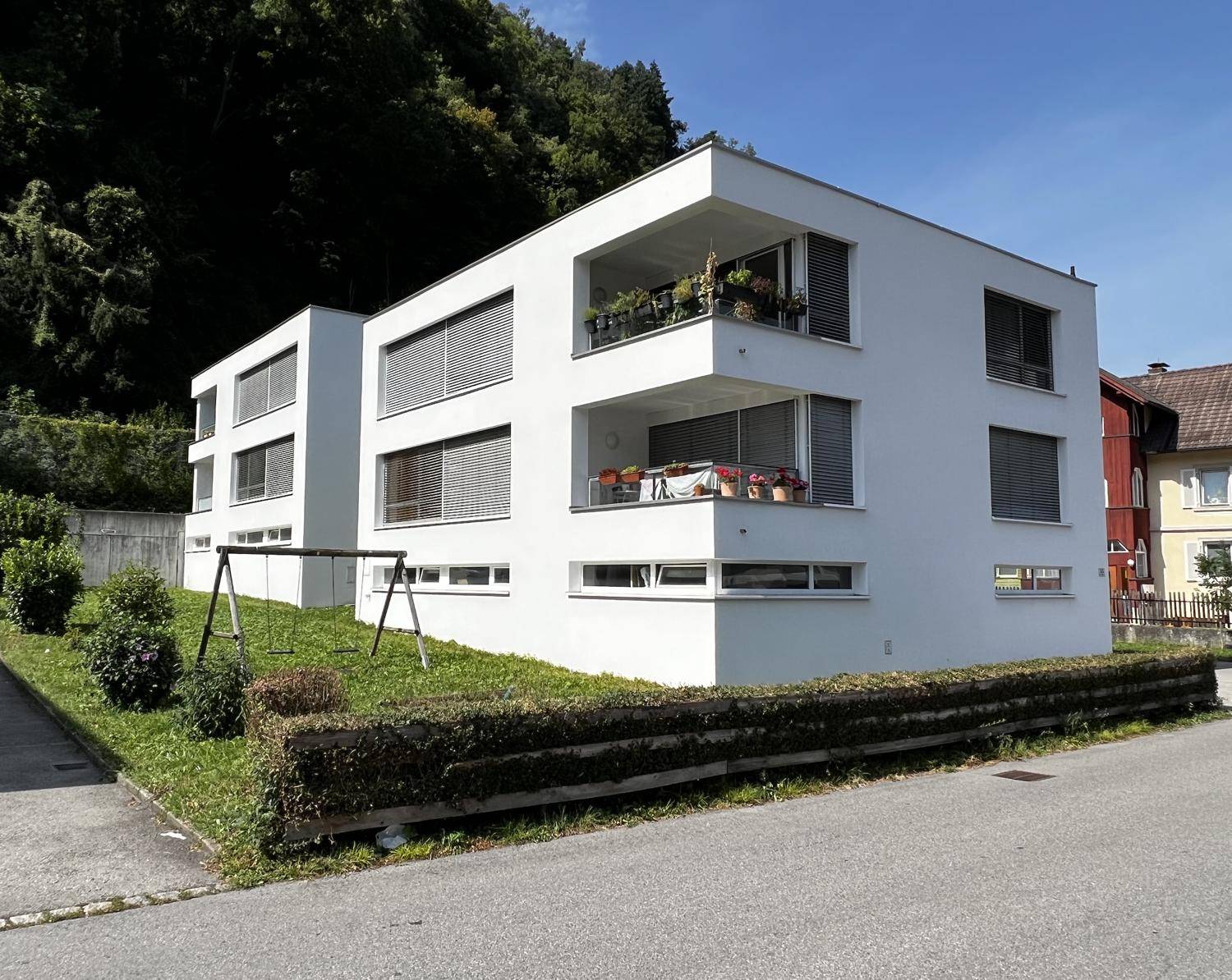 Modernes Wohngebäude mit weißen Fassaden, Balkonen und einem kleinen Spielbereich im Vorgarten.