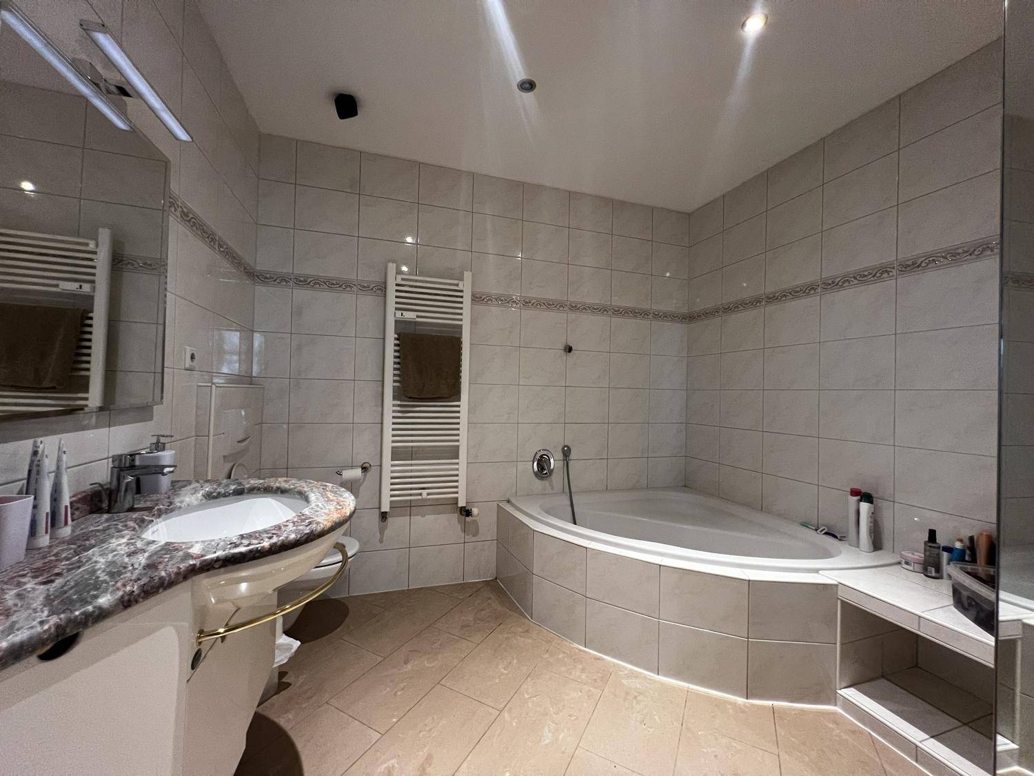 Badezimmer mit Eckbadewanne, Granitwaschtisch und hellen Fliesen, ausgestattet mit einem beheizten Handtuchhalter.