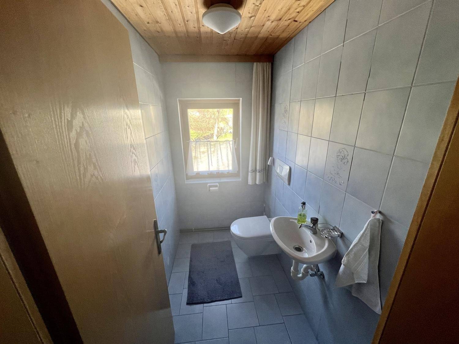 Kleines Gäste-WC mit Fenster, Wand-WC und Waschbecken in hellgrauen Fliesen.
