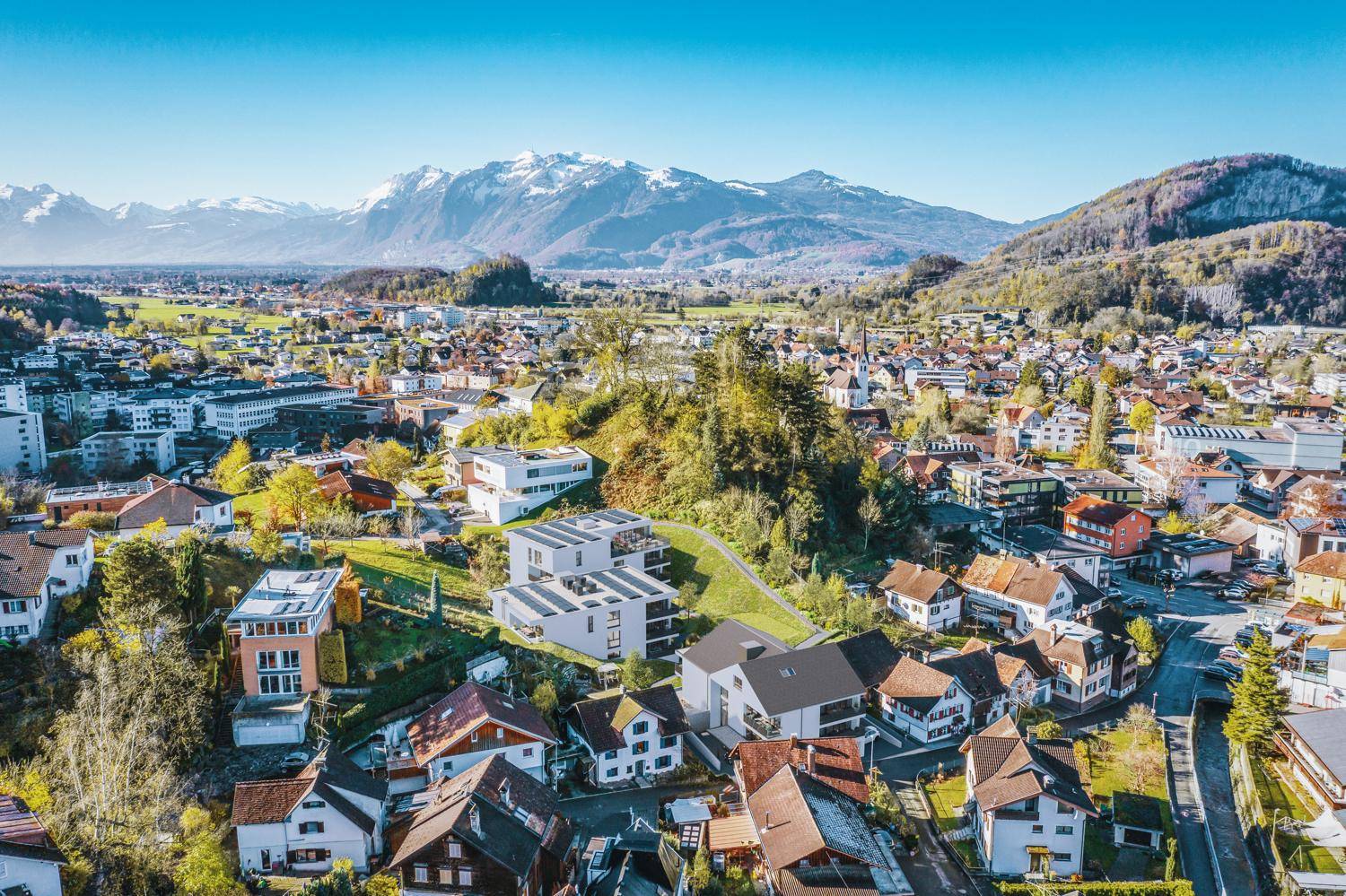 Panoramablick auf eine Wohnsiedlung mit modernen Häusern vor einer malerischen Bergkulisse unter blauem Himmel.
