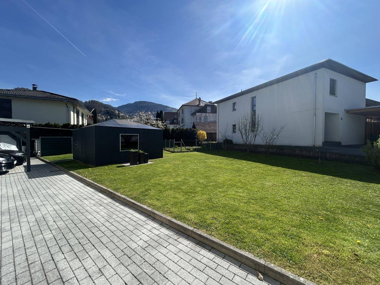 Gepflegter Garten mit Rasenfläche, gepflasterter Zufahrt und einem modernen Gartenhaus unter blauem Himmel.