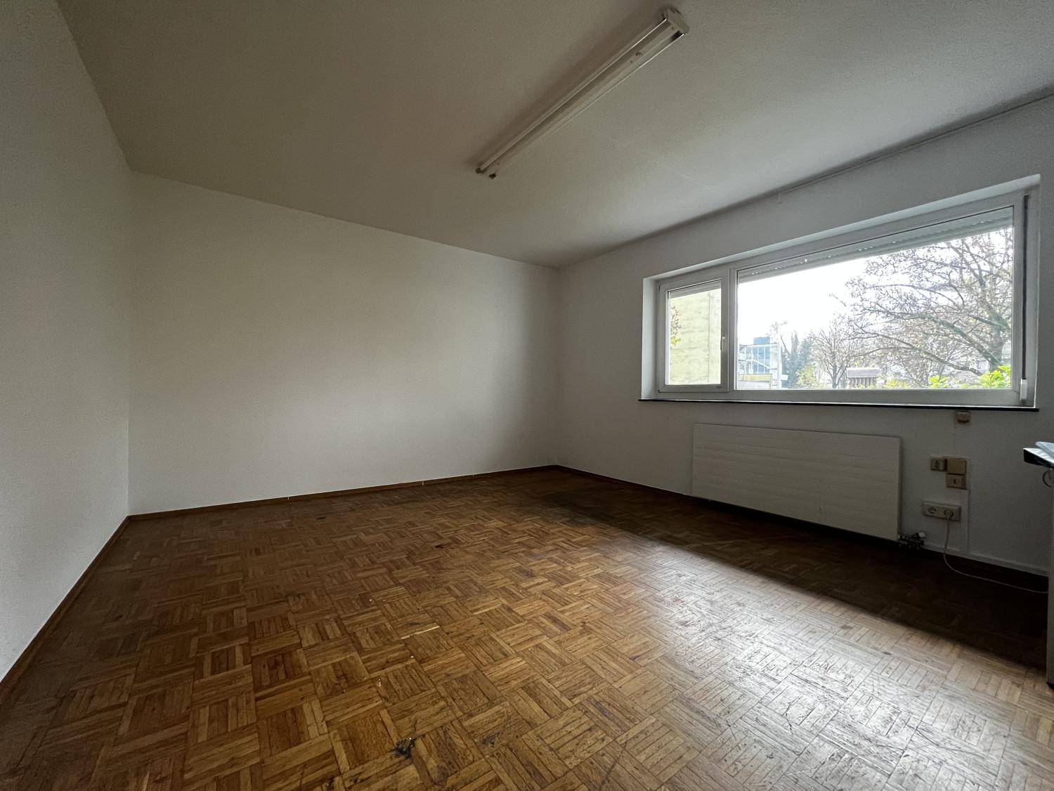 Leerer Raum mit Fischgrätparkettboden und großem Fenster, das viel Tageslicht hereinlässt.