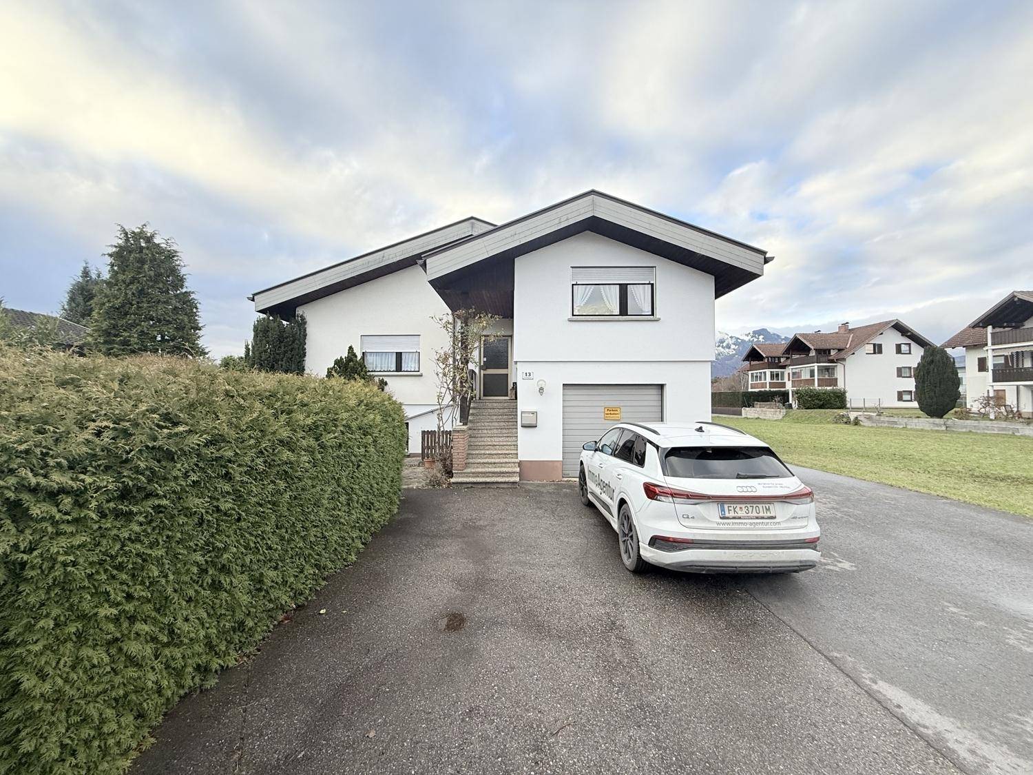 Das Einfamilienhaus von vorne mit Garage, Einfahrt und grüner Hecke, umgeben von Natur.