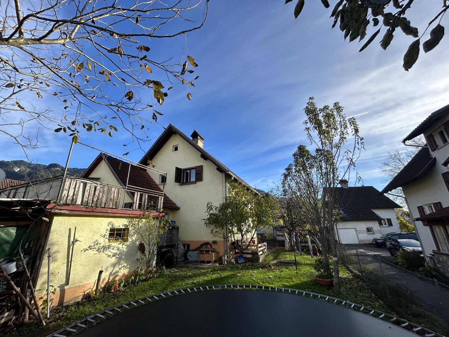 Hinteransicht des Hauses mit Garten, Trampolin und Blick auf die umliegende Landschaft unter blauem Himmel.