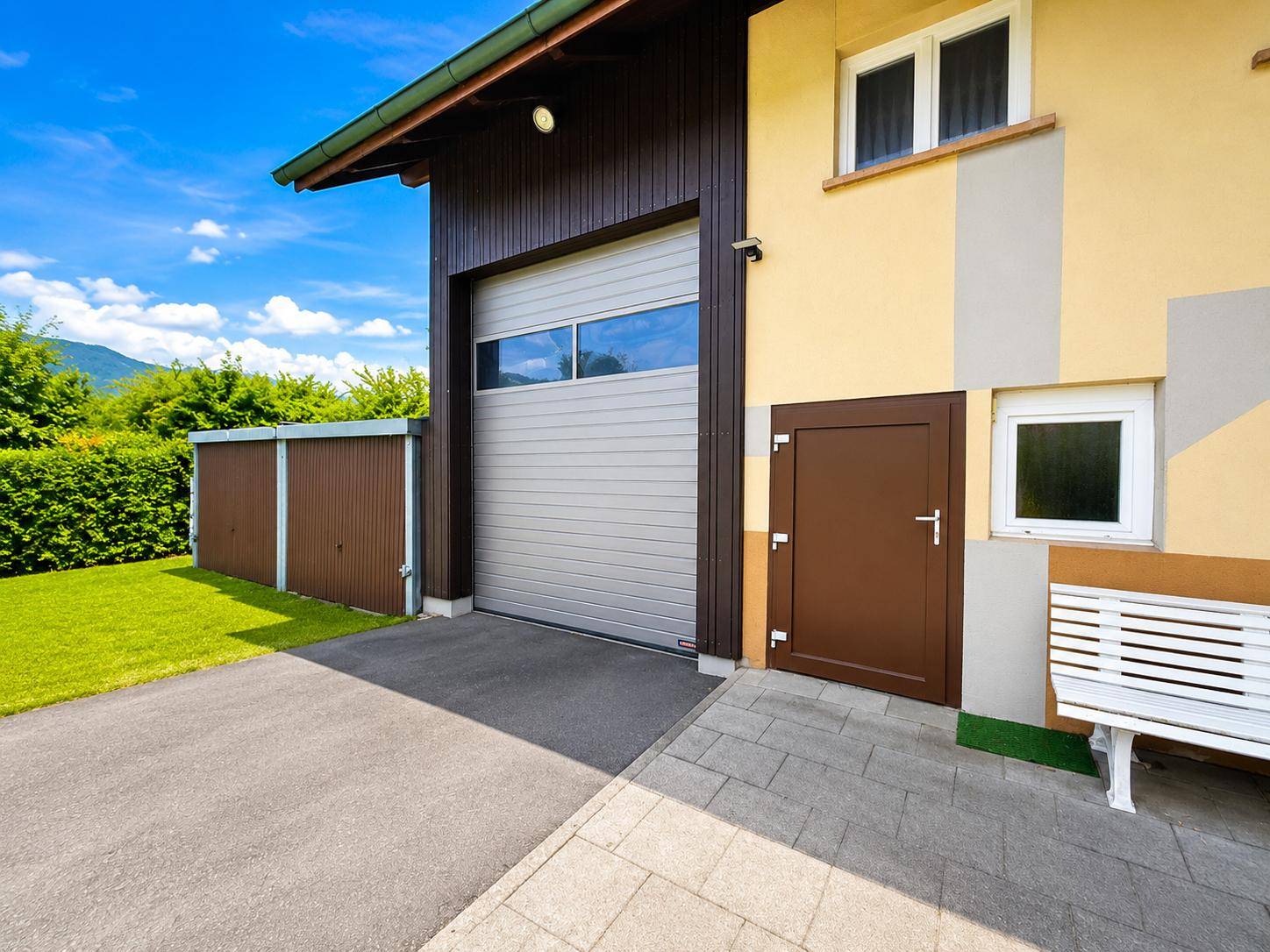 Garagentor und Nebeneingang des Hauses mit gepflastertem Bereich und kleiner Bank.