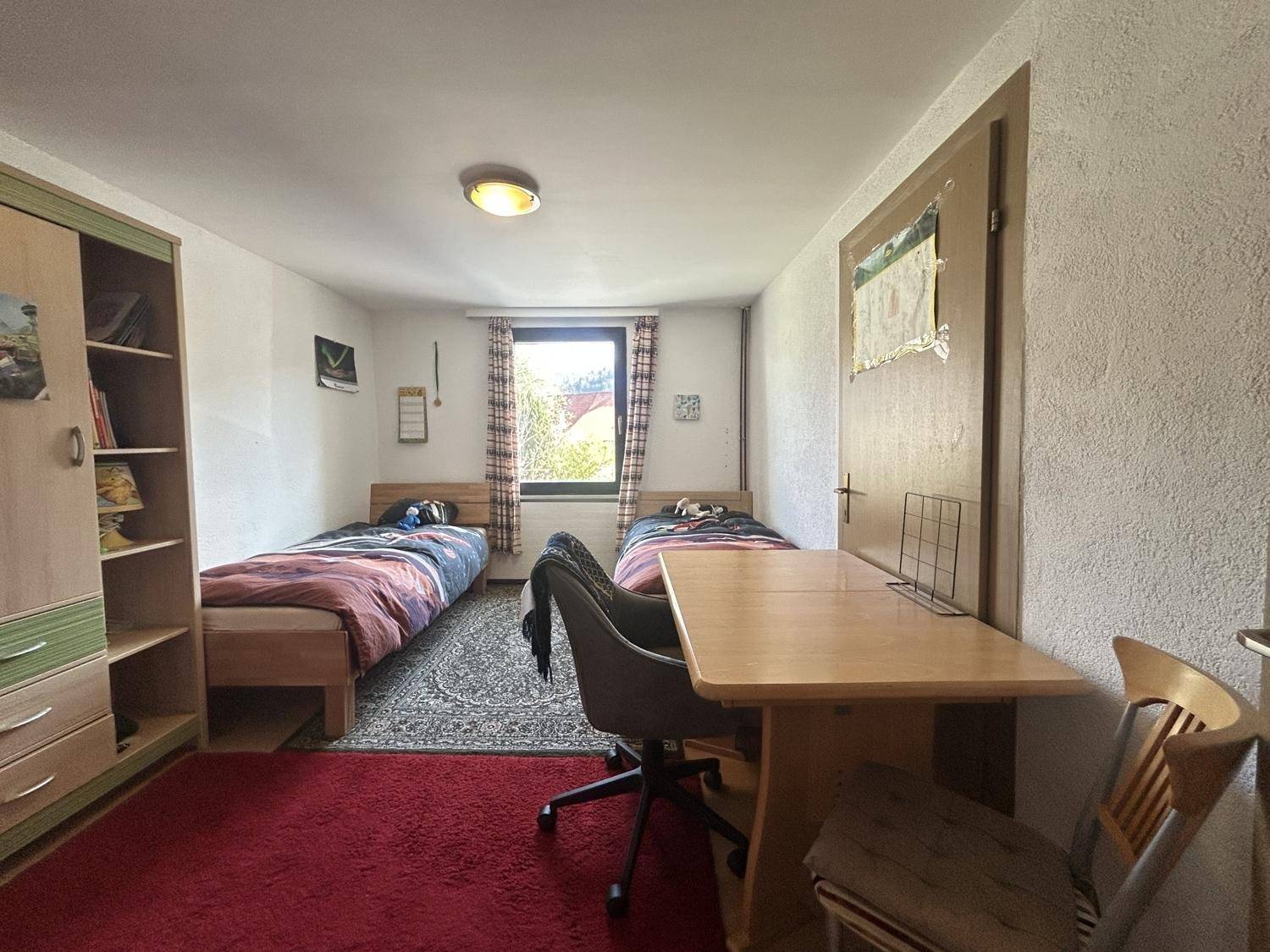 Kinderzimmer mit zwei Einzelbetten, Schreibtisch und rotem Teppich, gemütlich und funktional eingerichtet.