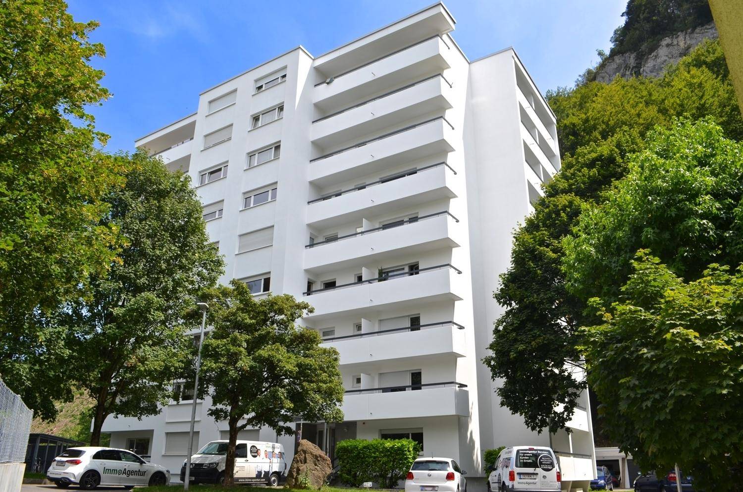 Modernes Mehrfamilienhaus mit weißer Fassade und zahlreichen Balkonen in grüner Umgebung.