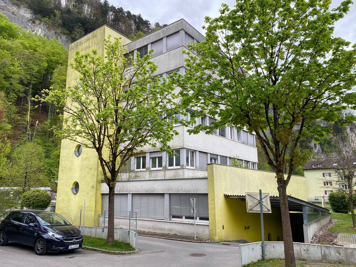 Modernes Wohngebäude mit gelber Fassade, Tiefgaragenzufahrt und Blick auf bewaldete Berge.