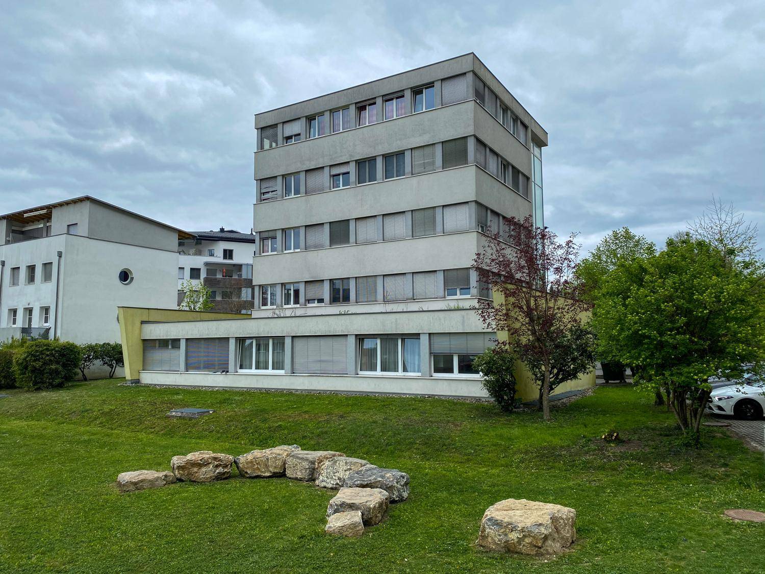 Mehrstöckiges Wohngebäude mit moderner Architektur und Grünfläche im Vordergrund unter bewölktem Himmel.