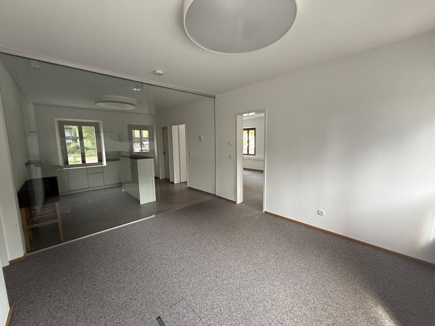 Offener Bürobereich mit Teppichboden und Blick auf eine moderne Küchenzeile hinter Glas.