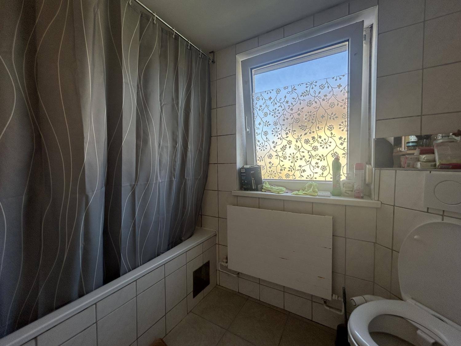 Gefliestes Badezimmer mit Badewanne, Duschvorhang und Fenster, funktional und hell eingerichtet.