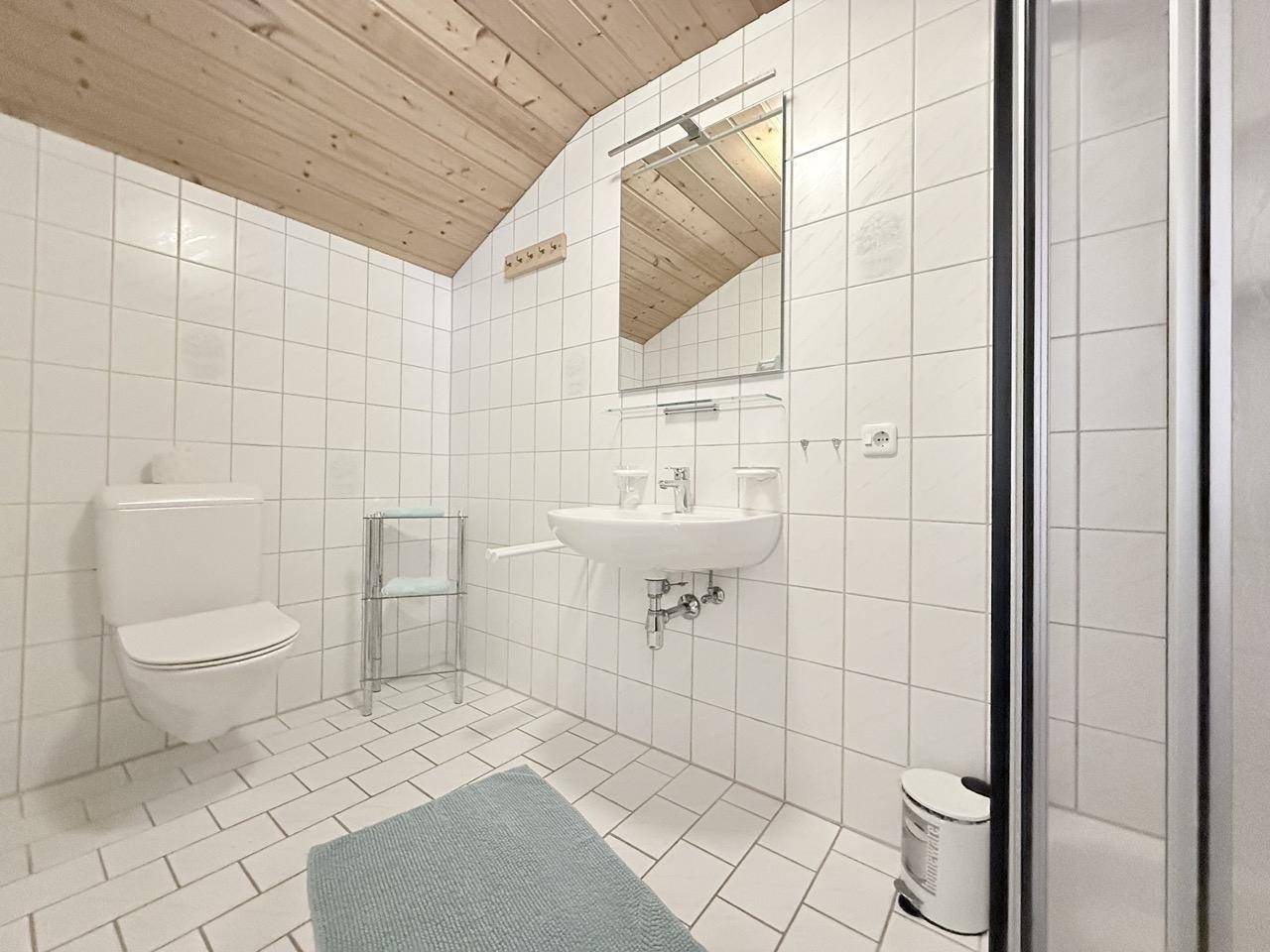 Helles Badezimmer mit weißen Fliesen, Waschbecken, Toilette und Duschbereich unter Dachschräge.