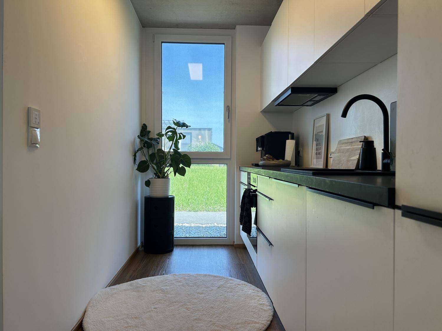 Schmale, moderne Küche mit weißen Schränken, dunkler Arbeitsplatte und Fenster zum Garten.
