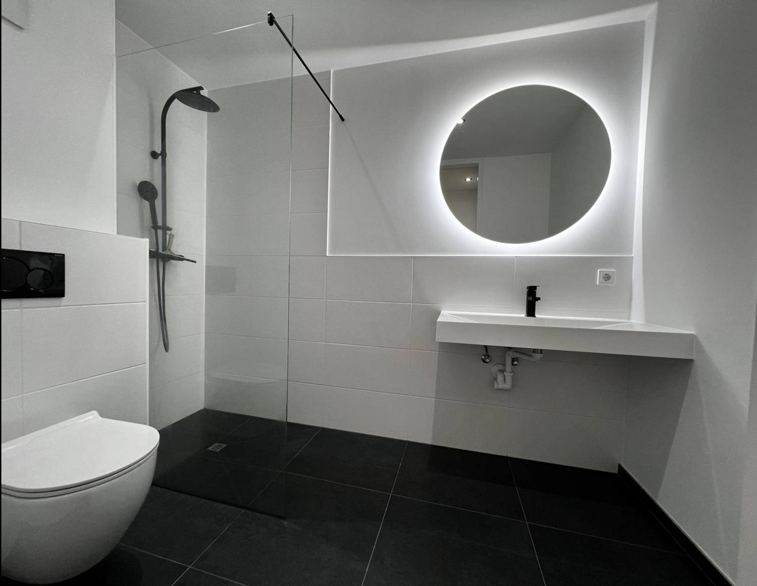 Modernes Badezimmer mit ebenerdiger Dusche, beleuchtetem Spiegel und dunklen Bodenfliesen.
