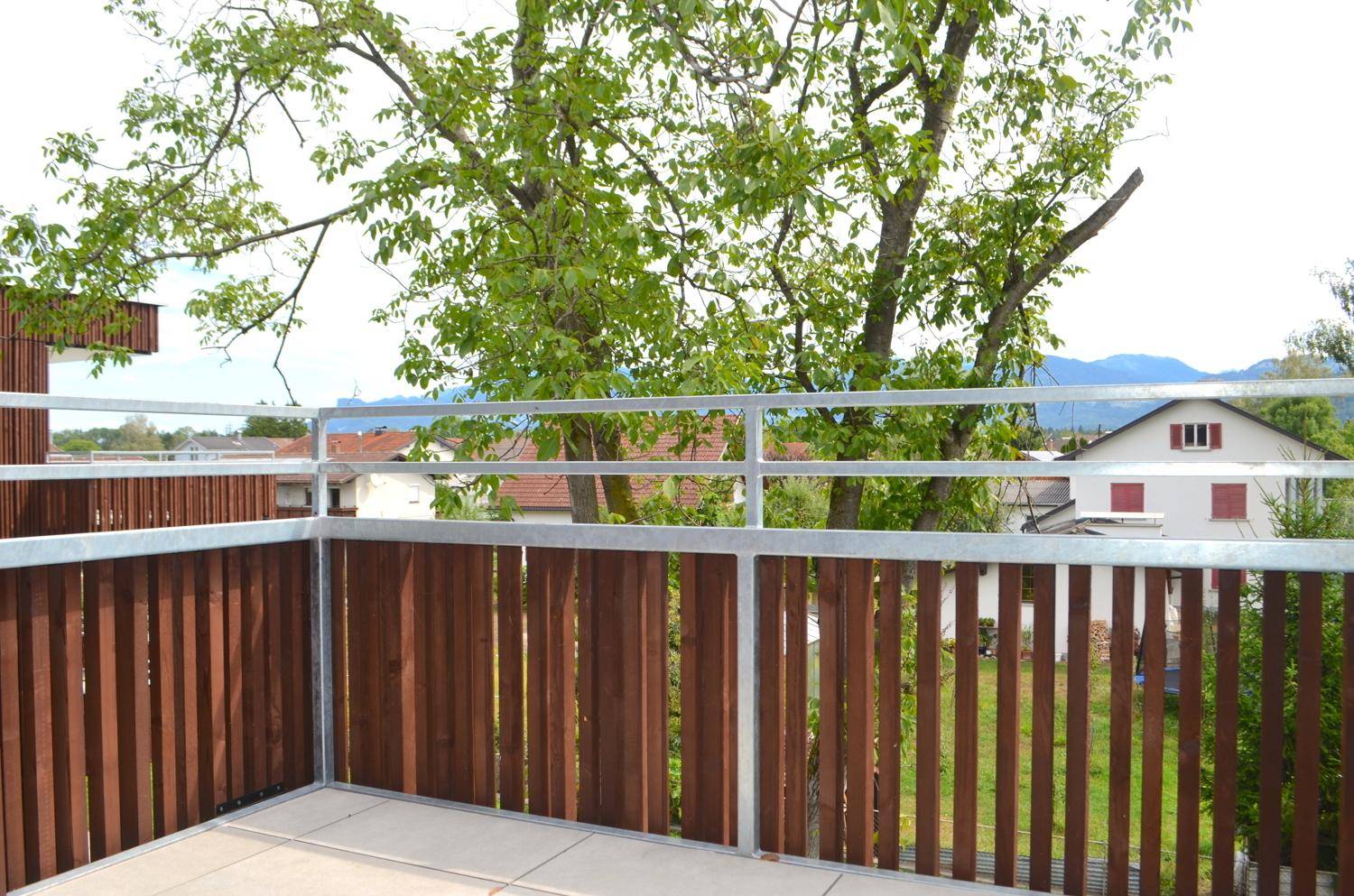 Balkon mit Holzelementen und Fliesenboden, bietet einen grünen Blick auf die Umgebung und Berge.