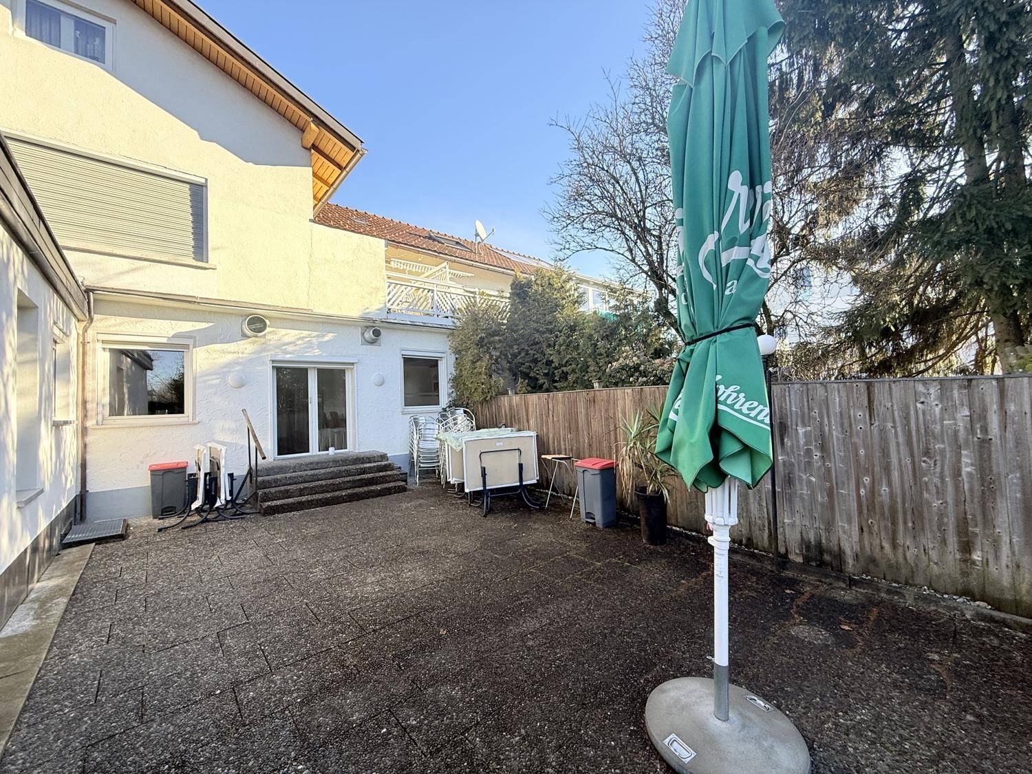 Hinterhof-Terrasse mit Sonnenschirm, gestapelten Stühlen und Zugang zum Gebäude.