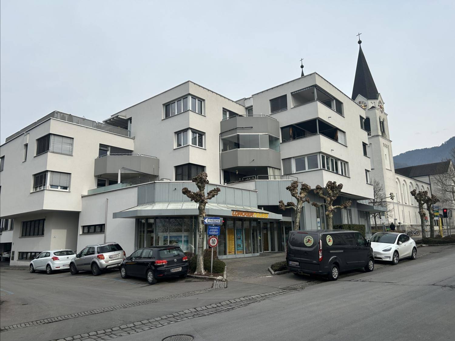 Ansicht eines modernen Wohn- und Geschäftshauses mit angrenzender Kirche und parkenden Autos.