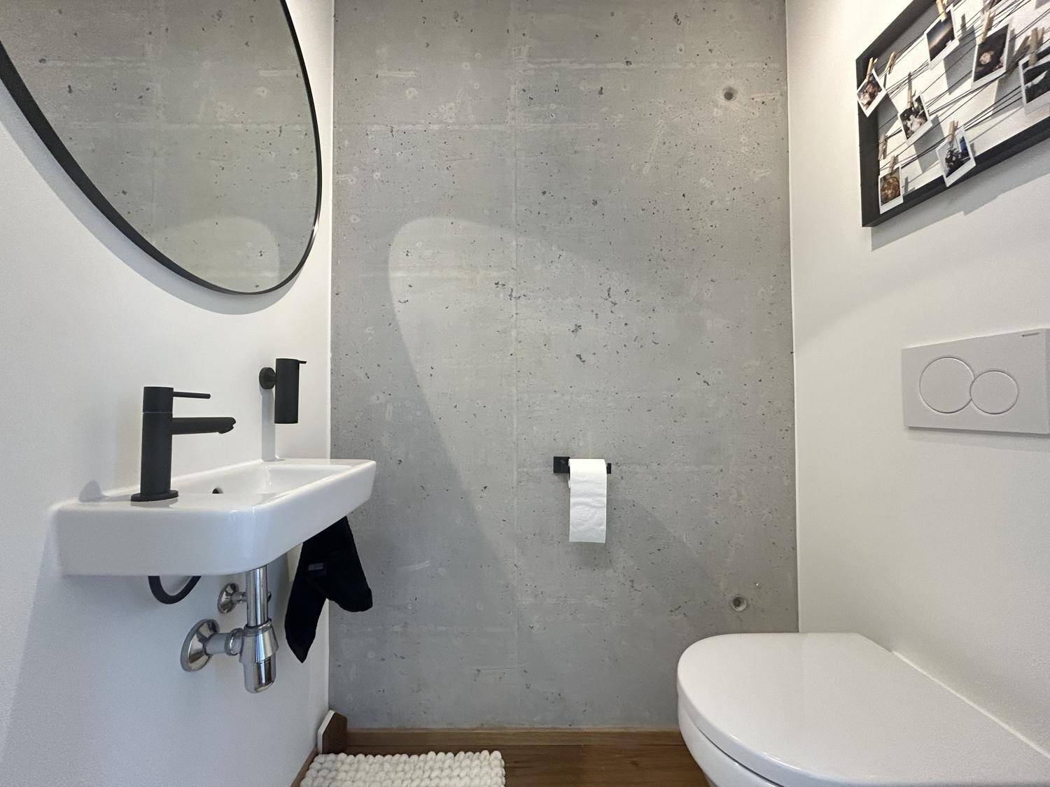 Modernes Gäste-WC mit schwebender Toilette, kleinem Waschbecken und Betonwand.
