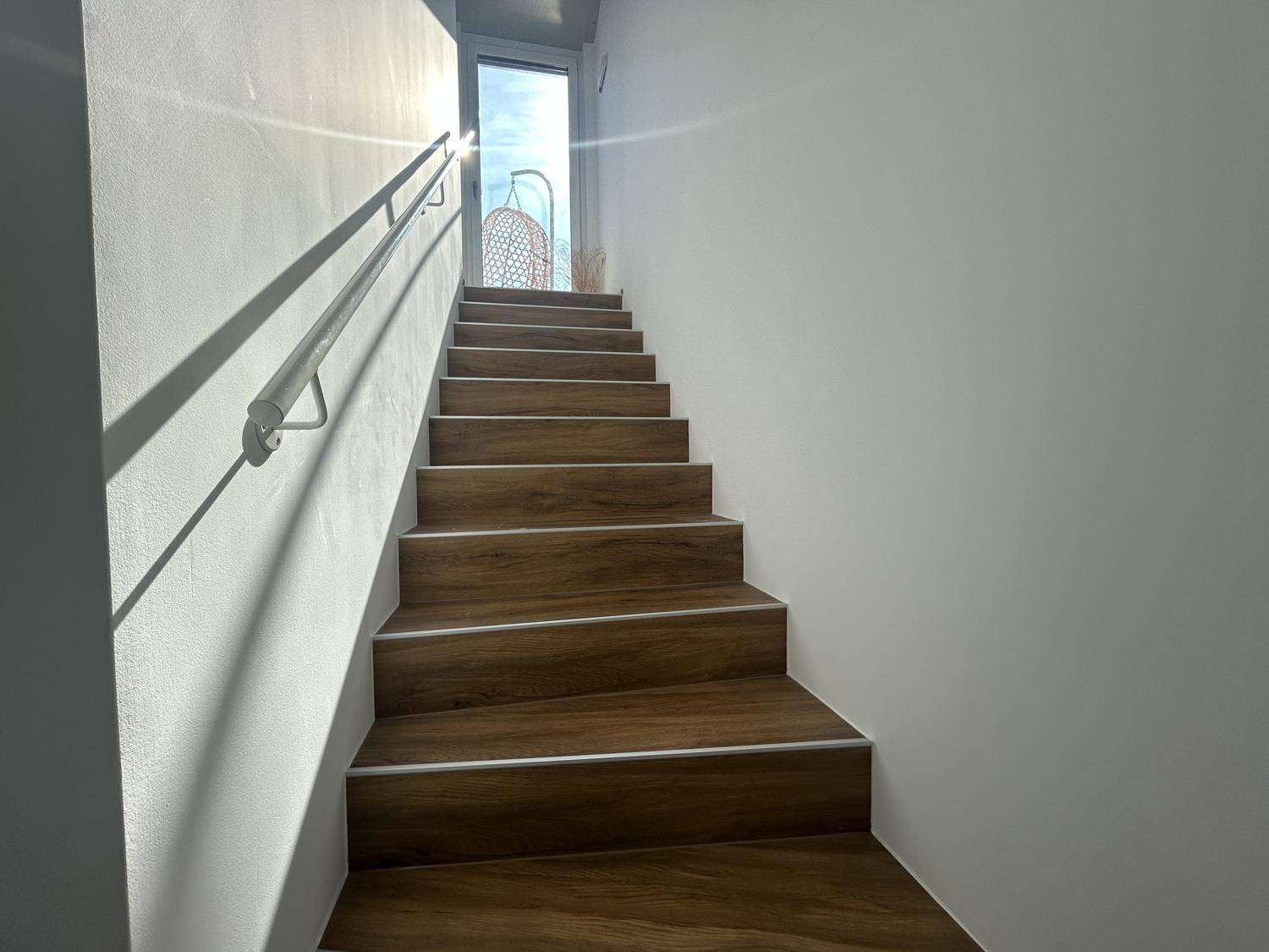 Helle Holztreppe mit Geländer, die zu einem sonnigen Außenbereich mit Terrasse führt.