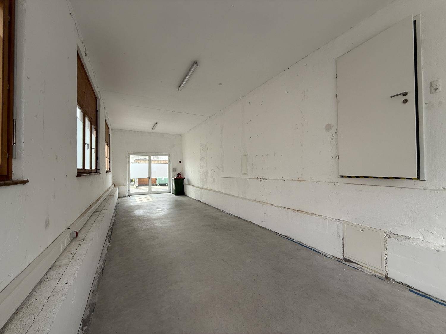 Langer Lagerraum mit Betonboden und Fenstern, der vielseitige Nutzungsmöglichkeiten bietet.