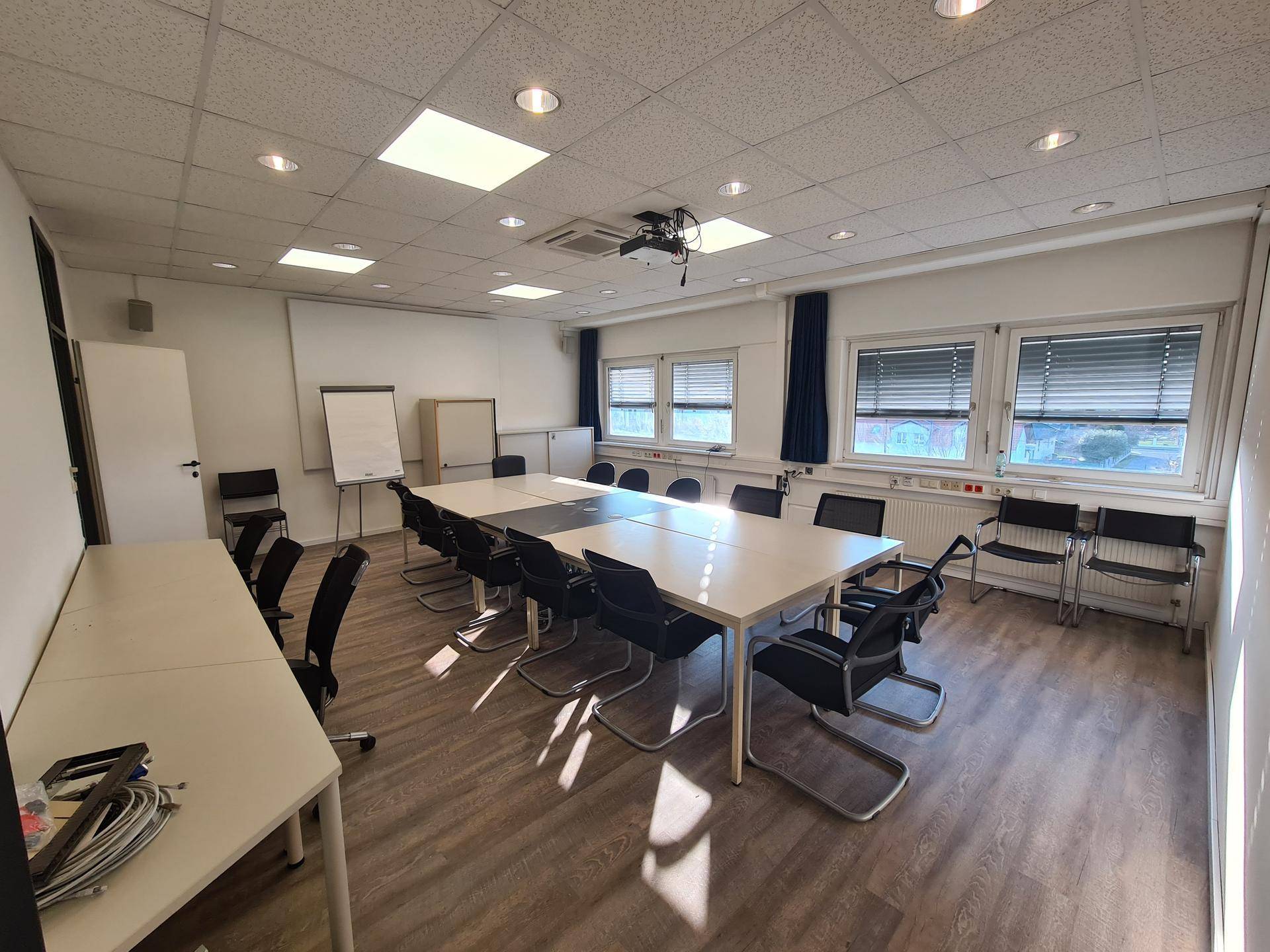 Großer Konferenzraum mit einem langen Tisch, vielen Stühlen, Beamer und Fenstern mit Jalousien.