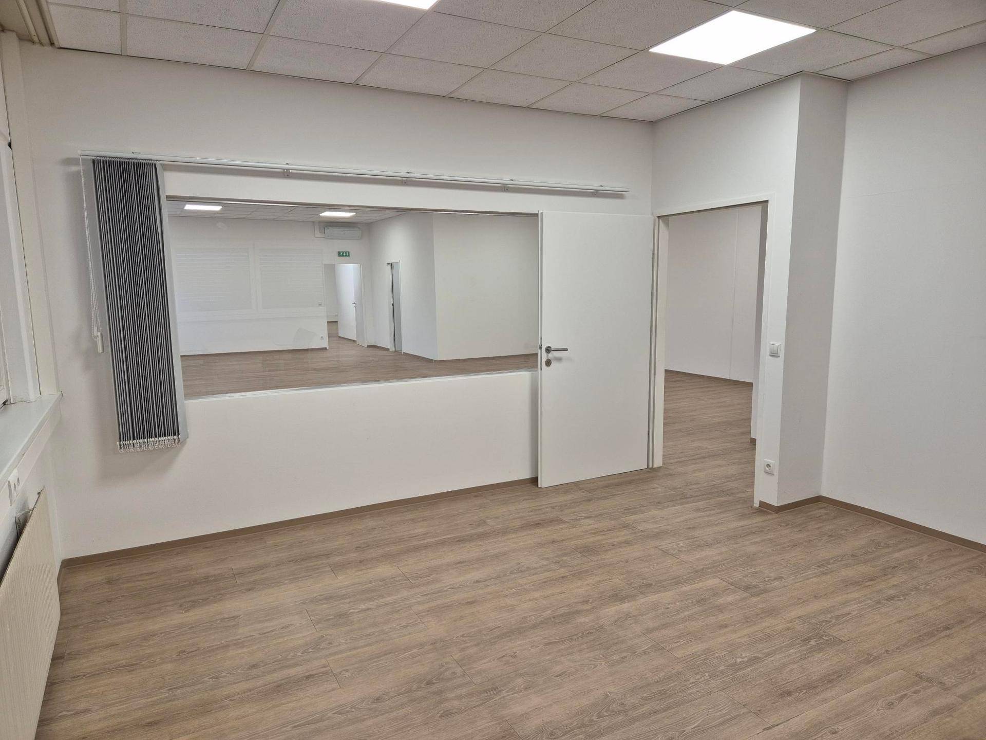 Heller Büroraum mit Laminatboden, weißen Wänden und einer großen Spiegelwand.