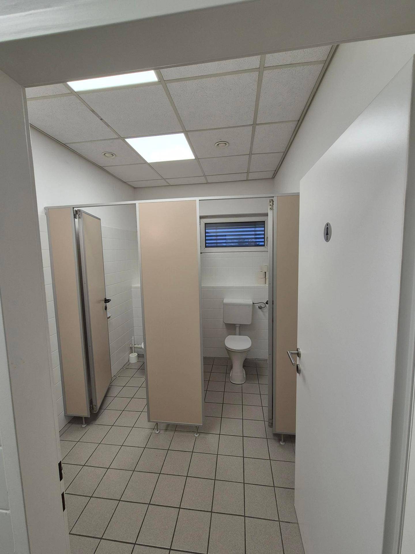 Sanitärbereich mit mehreren Toilettenkabinen, Fliesenboden und einem Fenster.