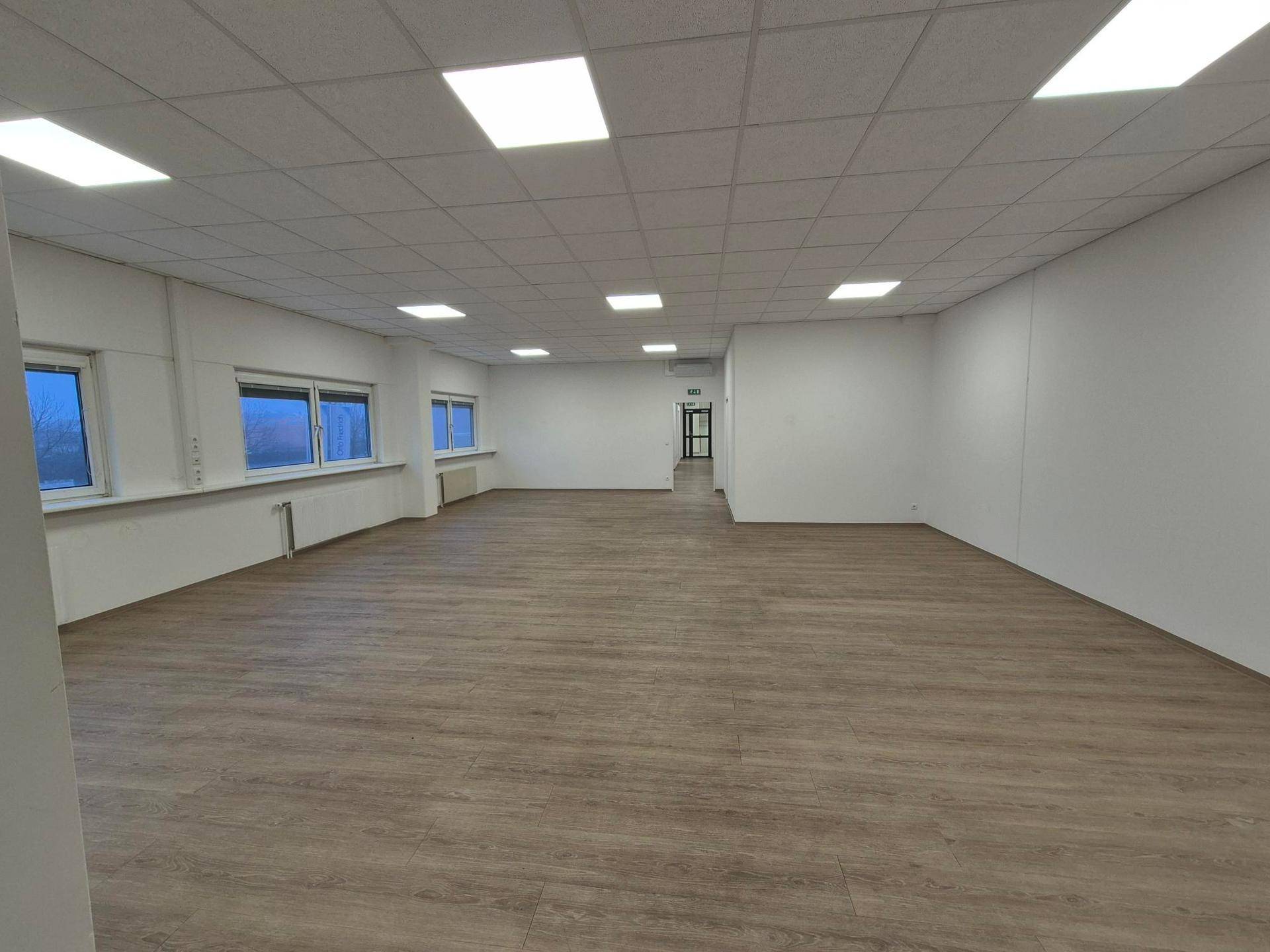 Großer, heller Büroraum mit Laminatboden, weißen Wänden und mehreren Fenstern.