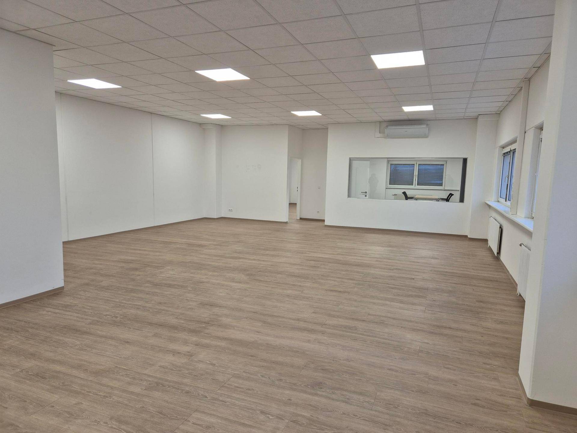 Geräumiger, leerer Büroraum mit Laminatboden, abgehängter Decke und großen Fenstern.