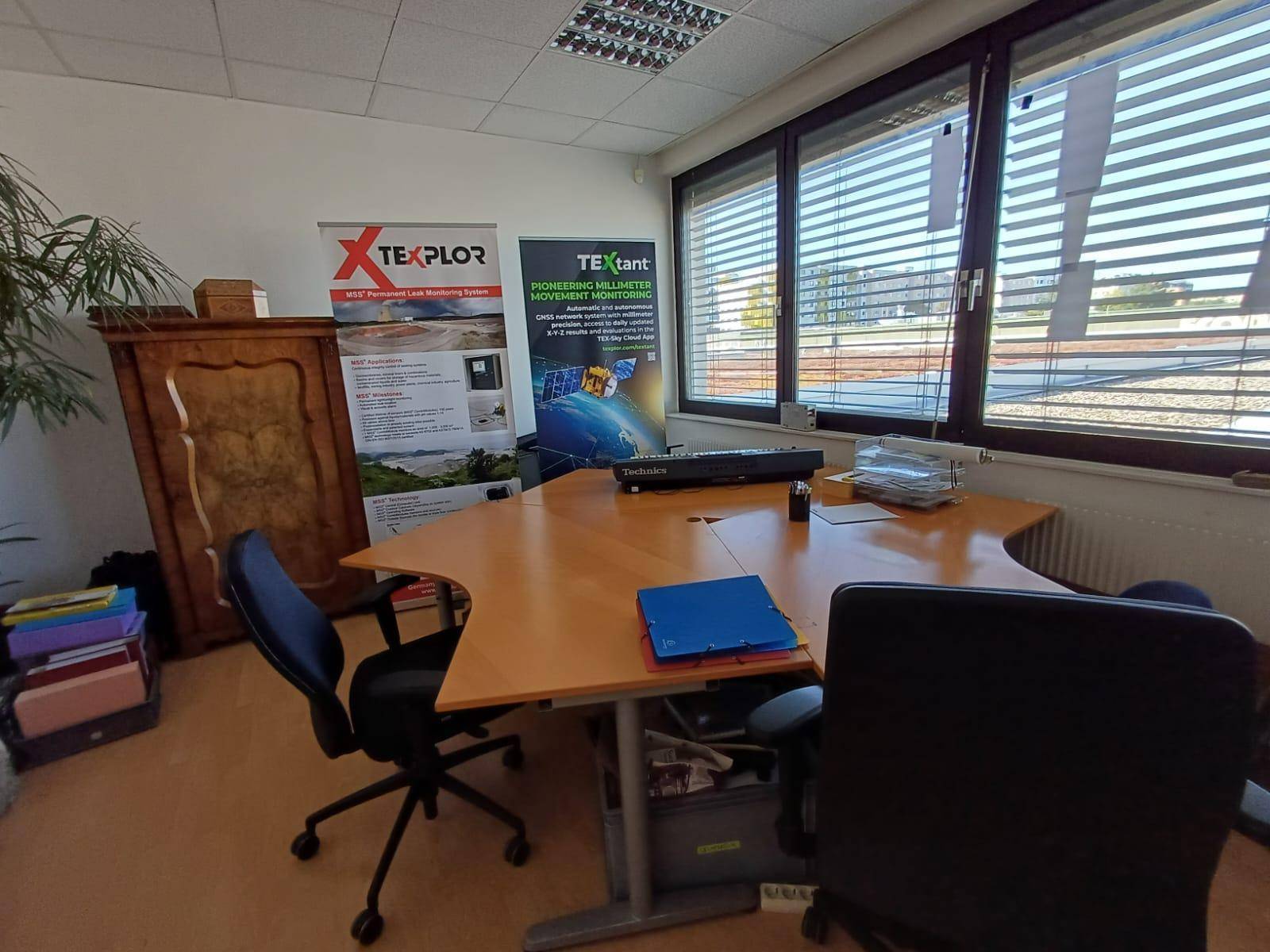 Büroraum mit Eckschreibtisch, Bürostühlen und Werbeplakaten an der Wand, gut beleuchtet.
