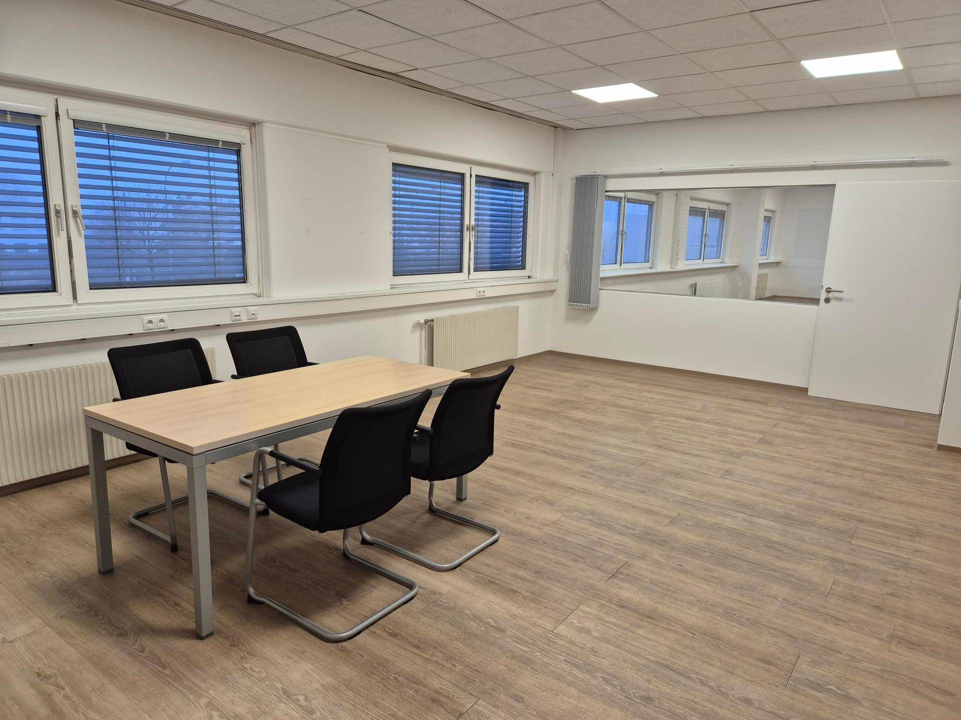 Heller Büroraum mit einem Tisch, Stühlen, Laminatboden und Fenstern mit Jalousien.