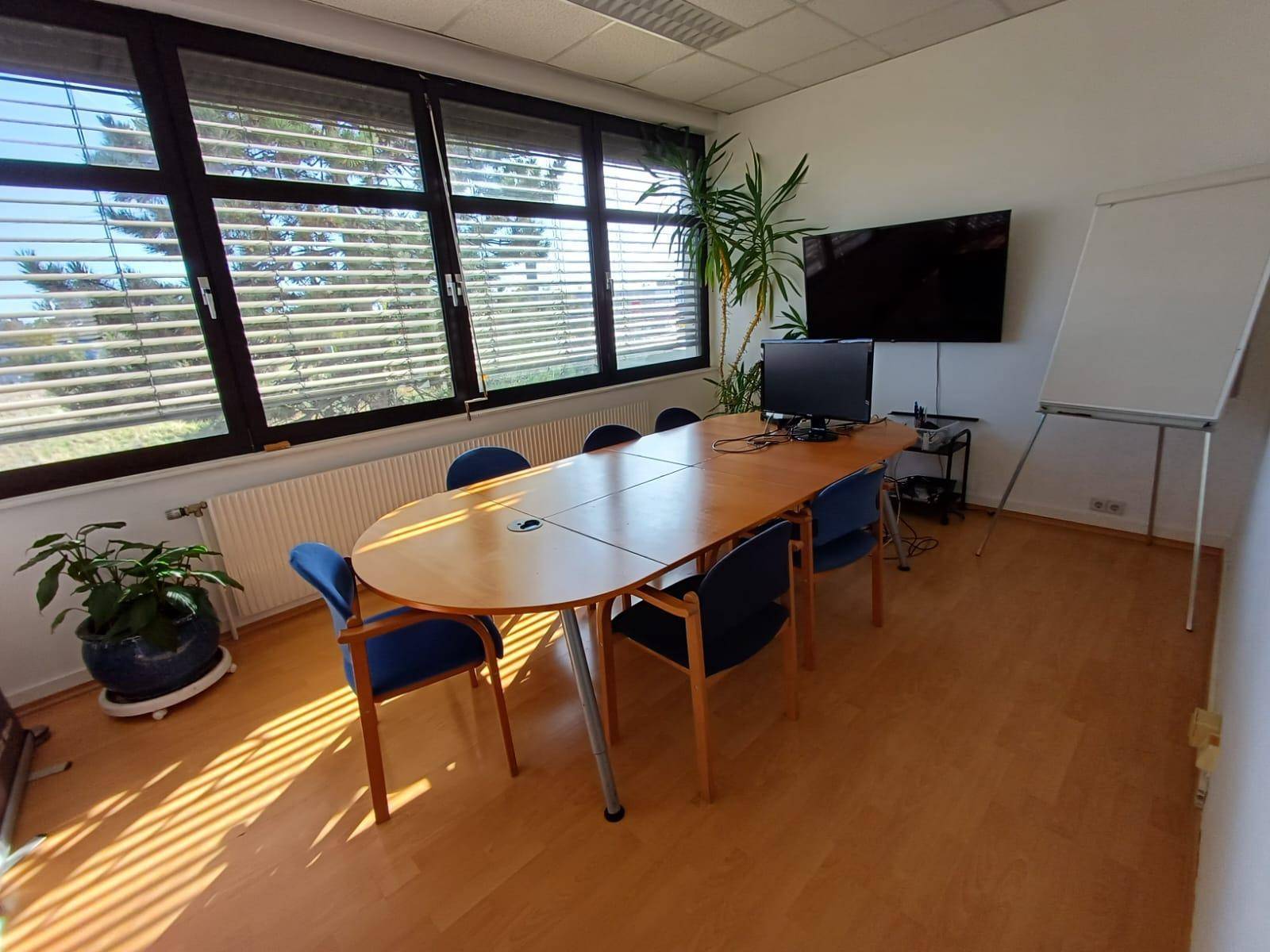 Heller Konferenzraum mit großem ovalen Tisch, Stühlen, Fernseher und großen Fenstern mit Jalousien.