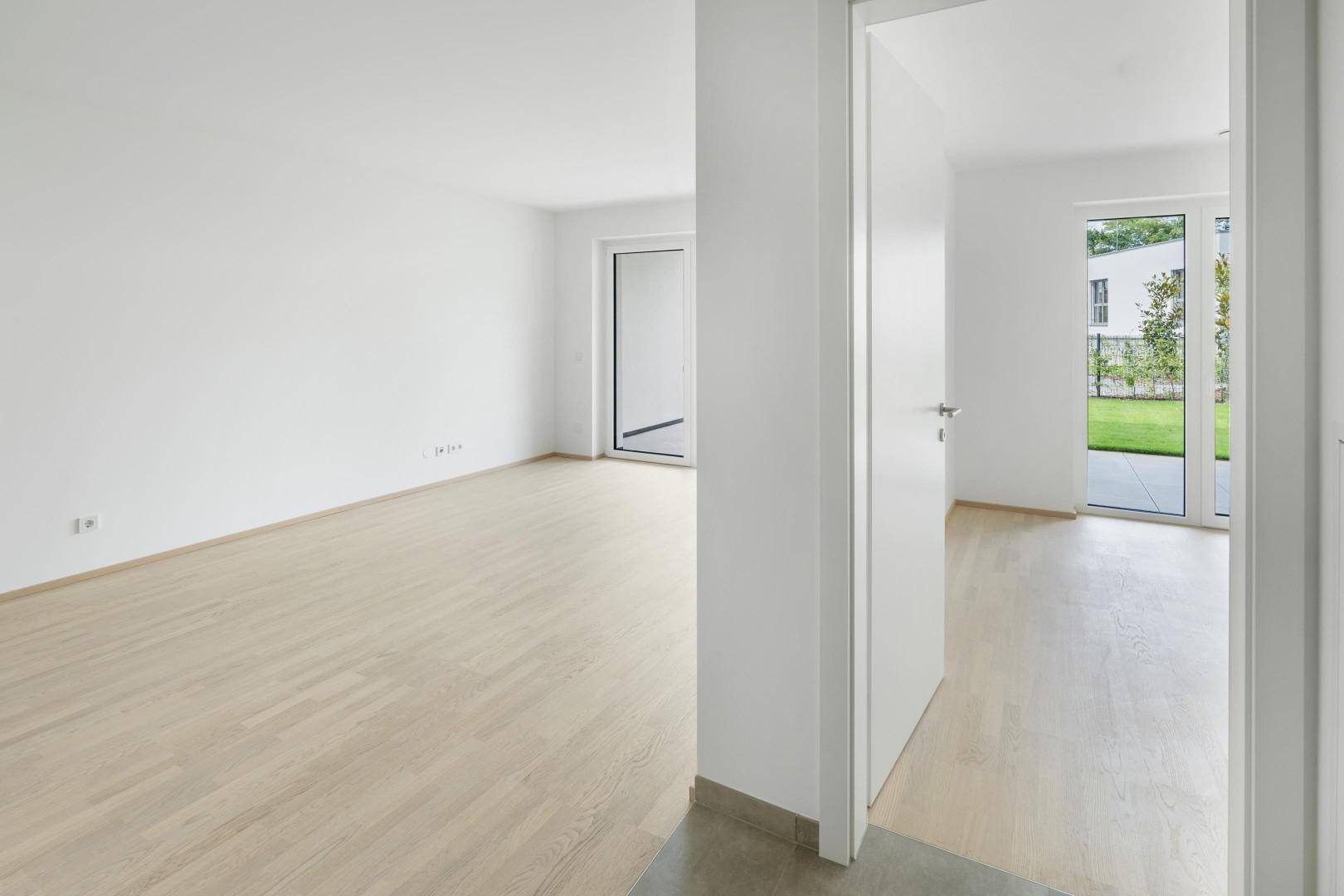 Offener Wohnbereich mit hellem Holzboden und Zugang zu einem weiteren Zimmer.